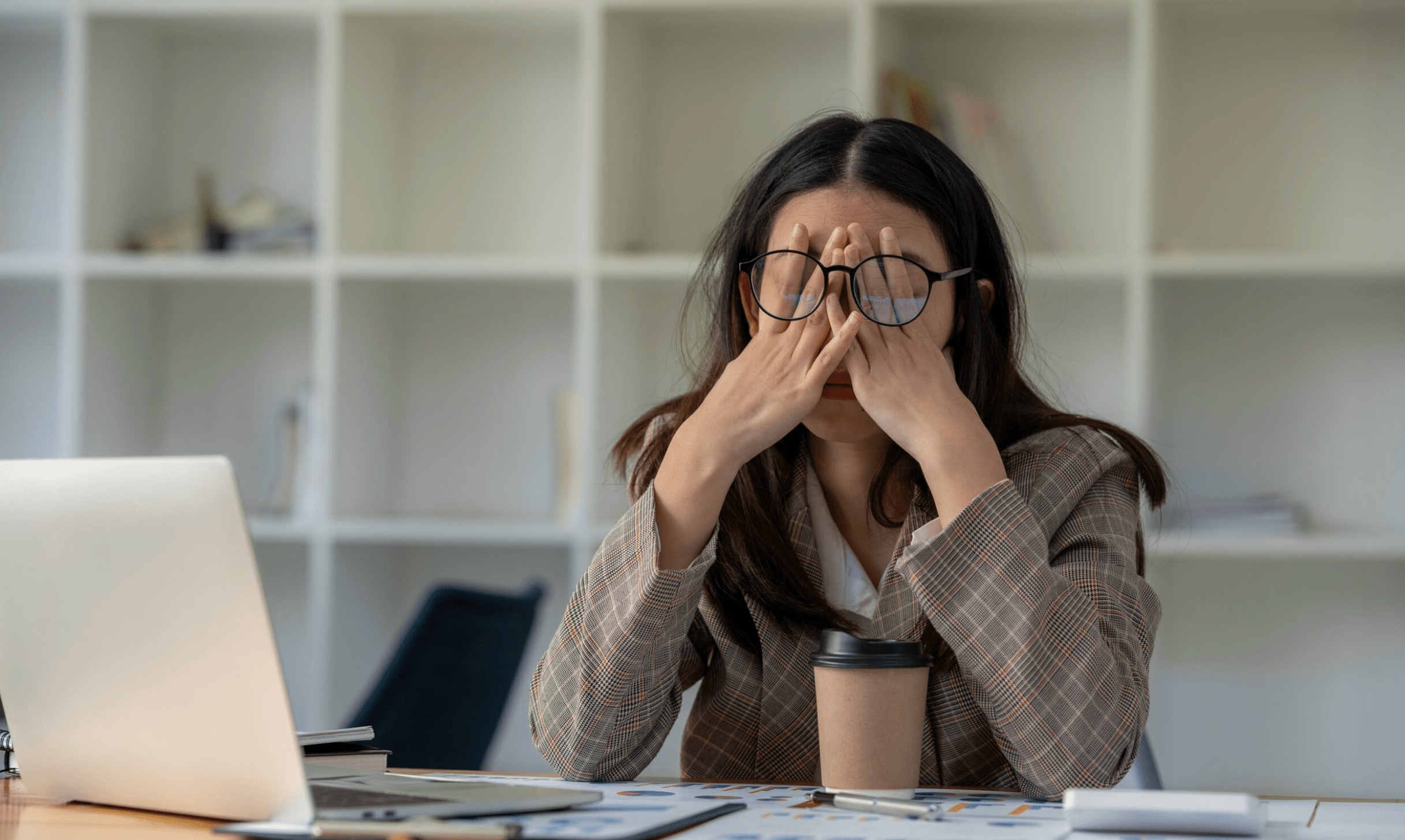 A young woman sitting at a desk with her laptop, pressing her fingers to her eyes from stress and fatigue. The image represents workplace stress and the need for natural ways to stay calm, such as CBD for stress and anxiety relief.